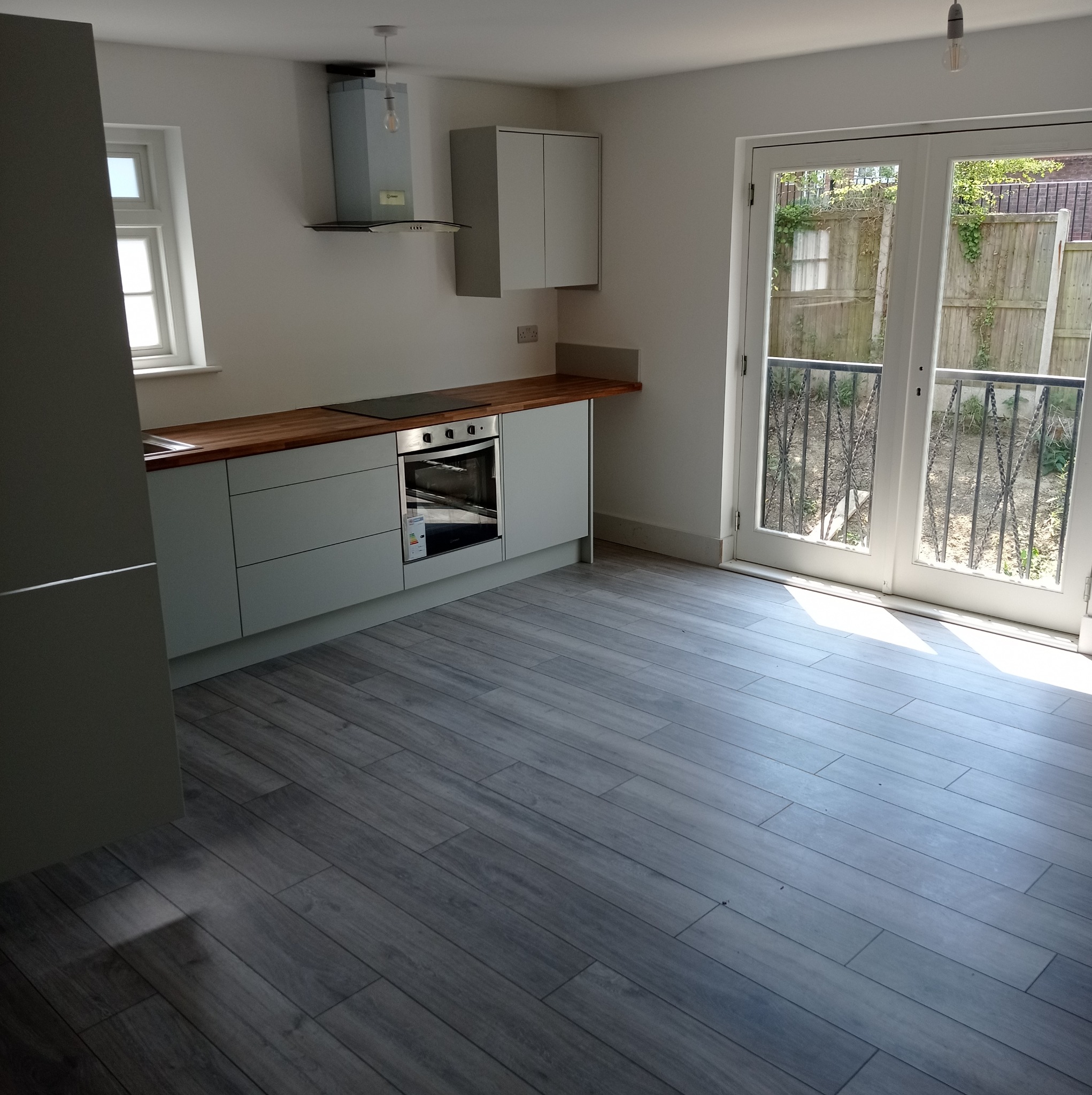 Jasna kuchnia z szarymi szafkami, drewnianym blatem i szarą podłogą z paneli, widok z niskiej perspektywy na wyjście balkonowe.