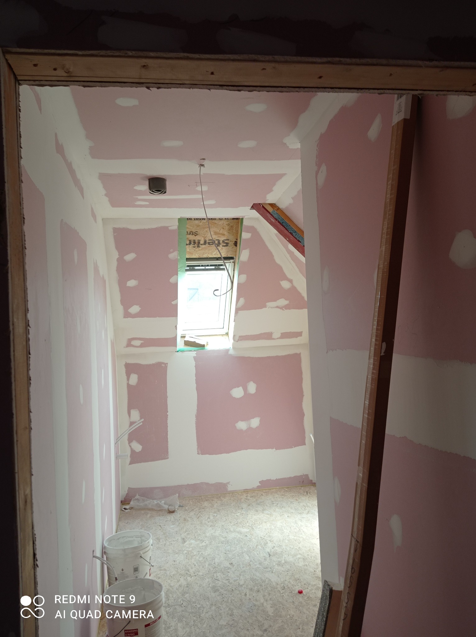 Poddasze w trakcie adaptacji: płyty gipsowo-kartonowe na ścianach i suficie, częściowo pokryte białą warstwą, okno dachowe z widoczną konstrukcją, dwa białe wiadra z farbą na podłodze z płyty OSB.