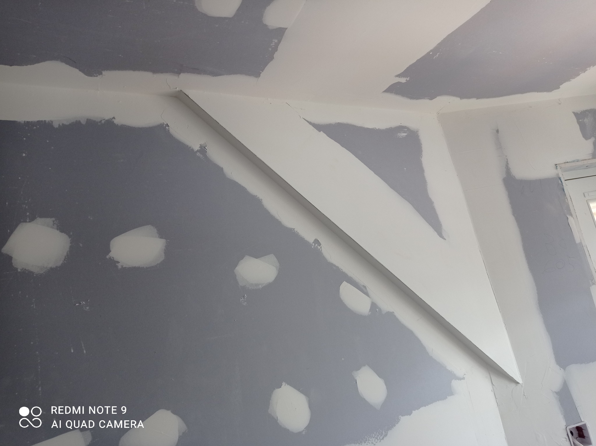 Ściana i sufit w trakcie prac wykończeniowych, widoczne fragmenty szpachli i biała listwa dekoracyjna pod sufitem, na ścianie widoczne punktowe naprawy szpachlą.