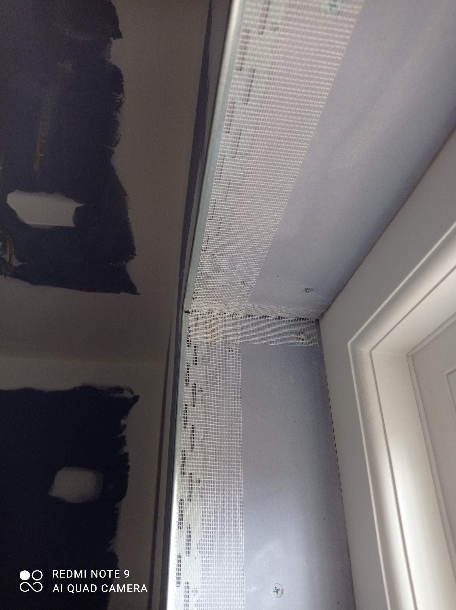 Przygotowanie do wyrównywania ściany przy ościeżnicy: siatka wzmacniająca nałożona na narożnik, widoczne ślady po wcześniejszych pracach na ścianie obok.