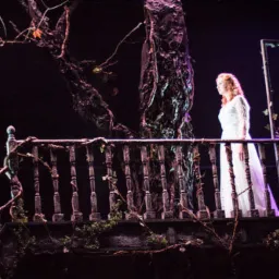 Scena teatralna z kobietą w białej sukni na balkonie, otoczona dekoracjami imitującymi drzewa i pnącza w mrocznej scenerii.