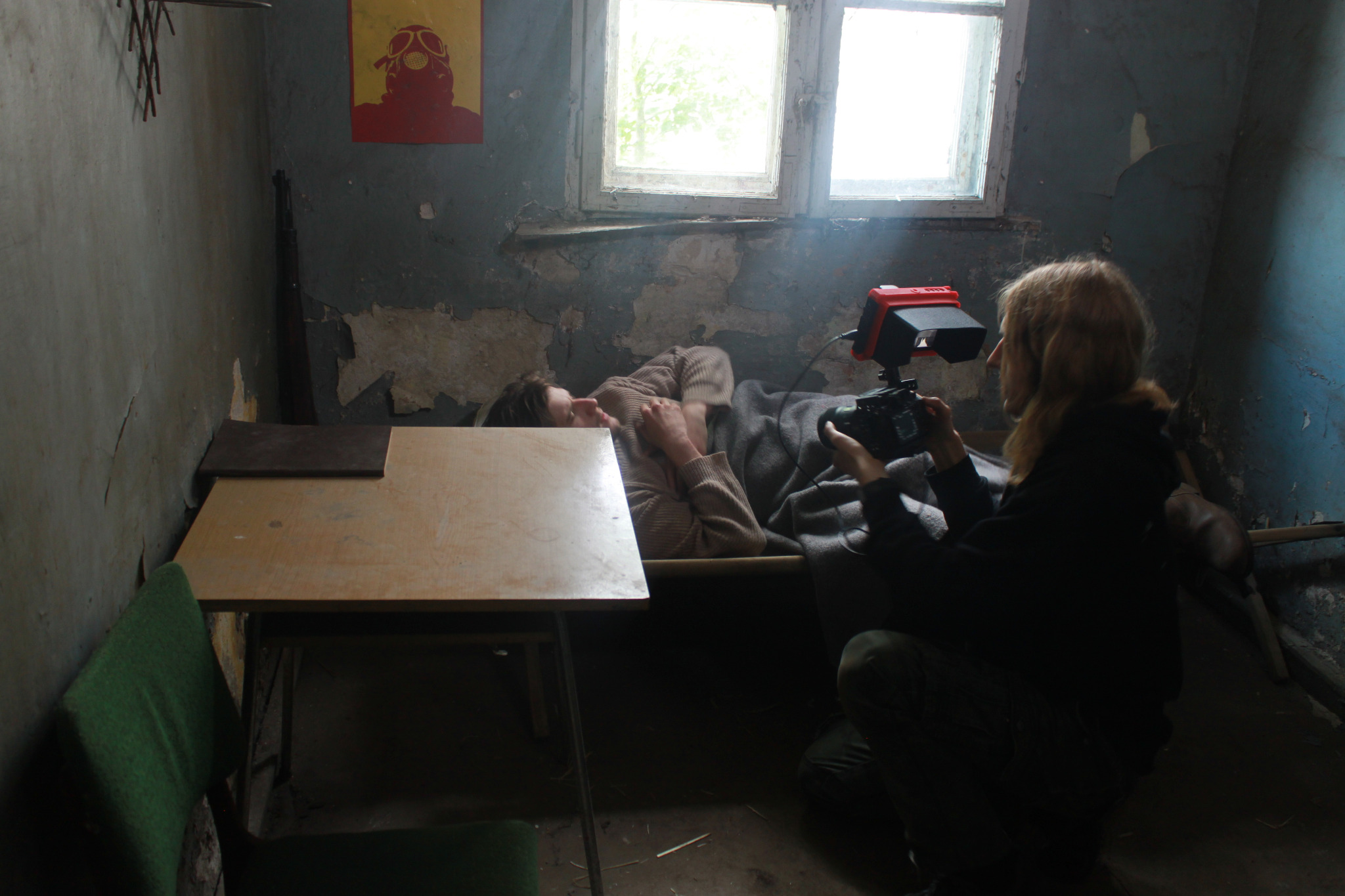 Ujęcie z planu filmowego w surowym wnętrzu: osoba filmuje leżącego aktora z użyciem kamery z lampą, widoczny stolik i plakat na ścianie.