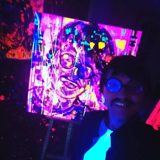 Mężczyzna w okularach pozuje na tle fluorescencyjnego portretu na sztaludze, oświetlonego światłem UV.