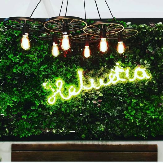 Oryginalna lampa z żarówkami Edisona zawieszona nad neonowym napisem na ścianie z zielonych roślin.