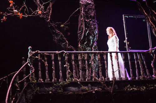 Scena teatralna z kobietą w białej sukni na balkonie, otoczona dekoracjami imitującymi drzewa i pnącza w mrocznej scenerii.