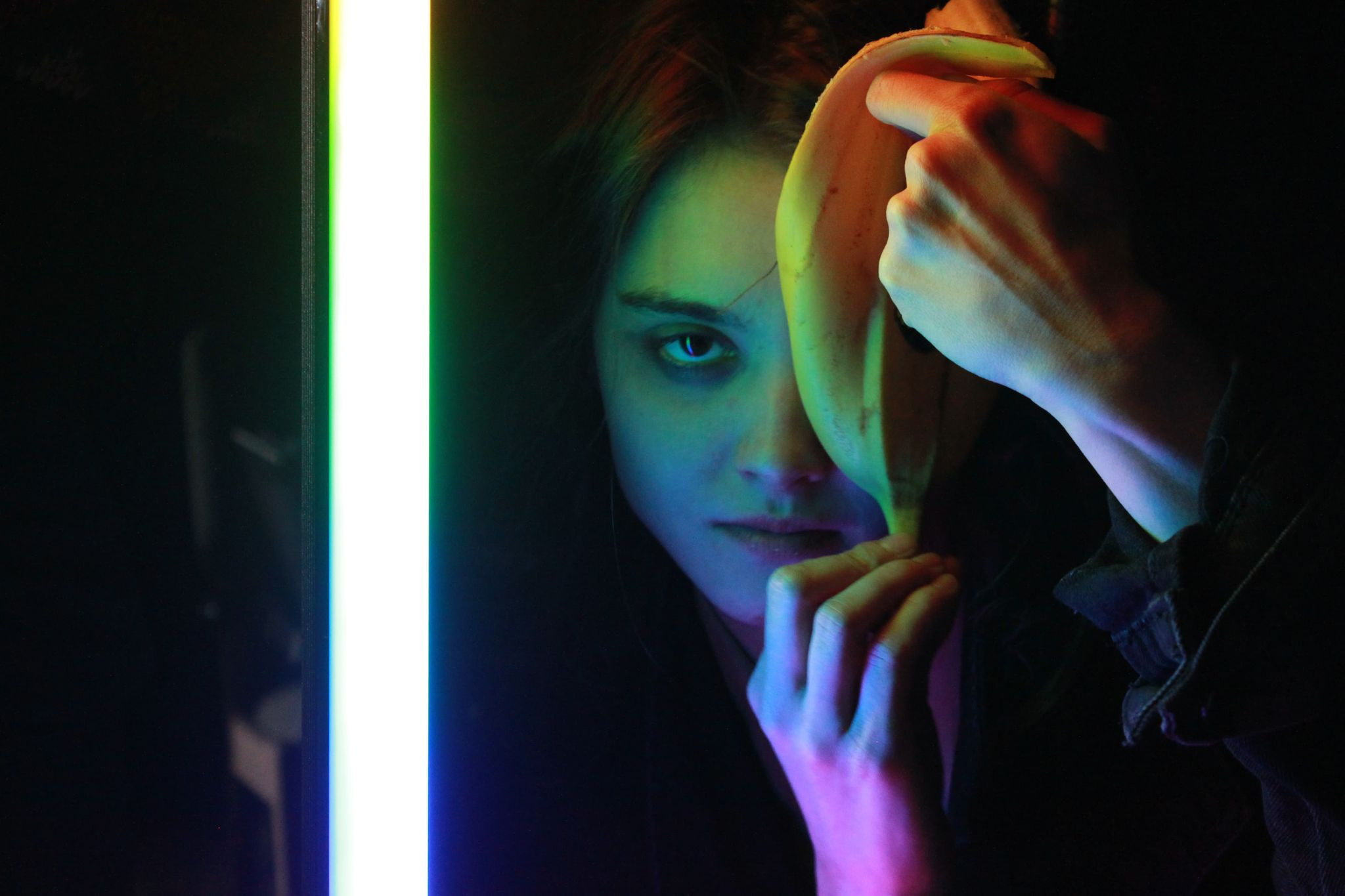 Portret młodej kobiety z bananem w dłoni, oświetlonej neonowym światłem w odcieniach zieleni, niebieskiego i różu, z intensywnym spojrzeniem w obiektyw.