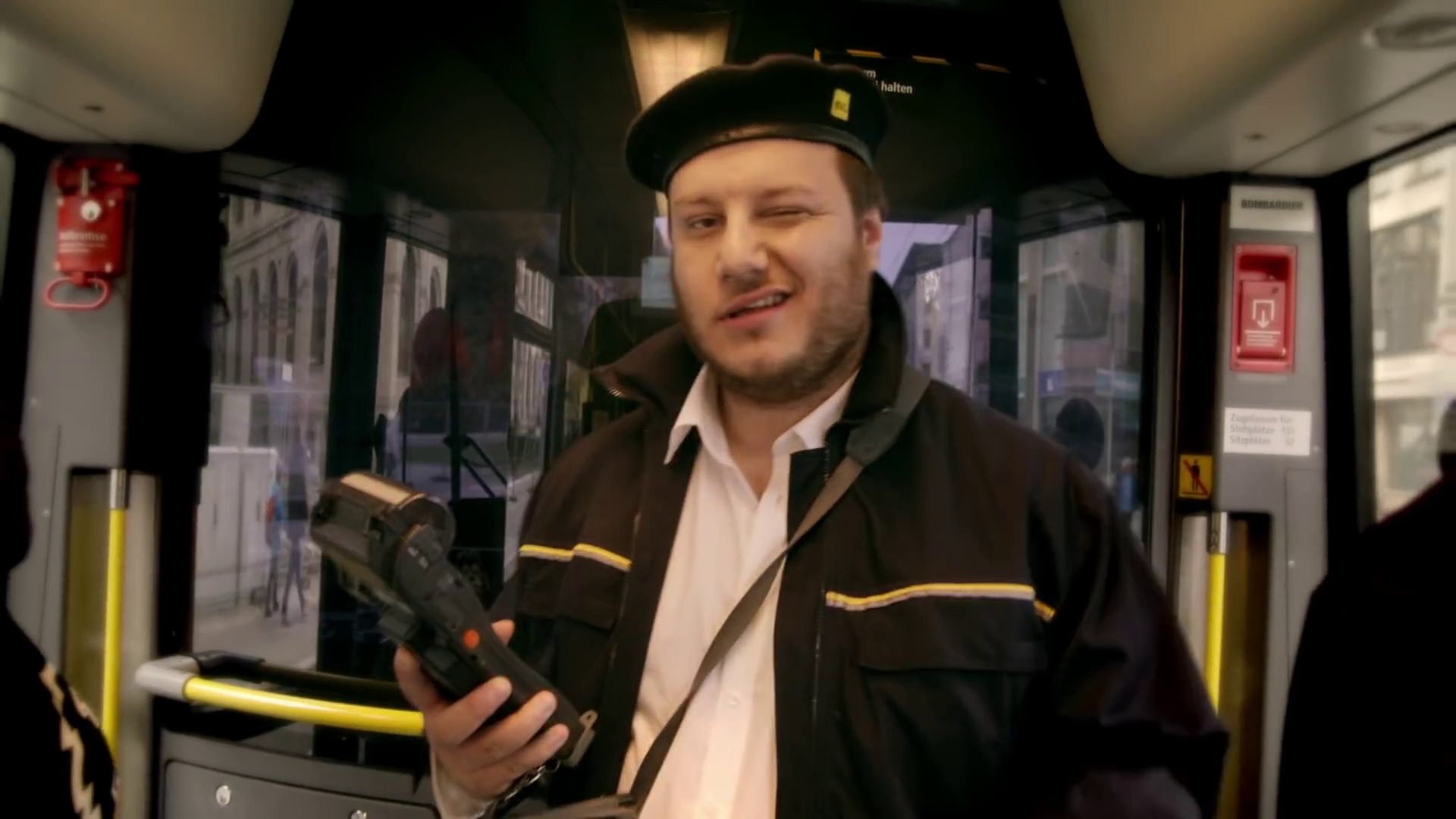 Mrugający mężczyzna w uniformie, prawdopodobnie kontroler biletów, w autobusie w Bydgoszczy, trzymający elektroniczny czytnik biletów.