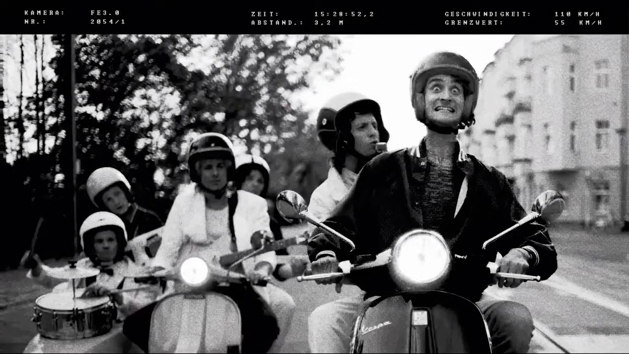 Czarno-białe zdjęcie grupy osób na skuterach Vespa, z przodu mężczyzna z szeroko otwartymi oczami, za nim inni uczestnicy, w tle budynki miejskie, u góry parametry kamery i prędkości.