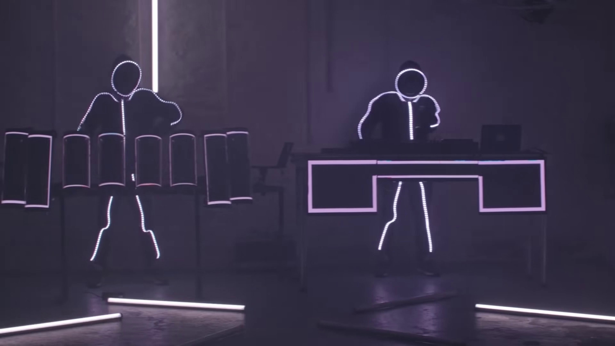 Dwóch performerów w podświetlanych strojach grających na elektronicznych instrumentach w ciemnym pomieszczeniu, z dodatkowym oświetleniem neonowym.