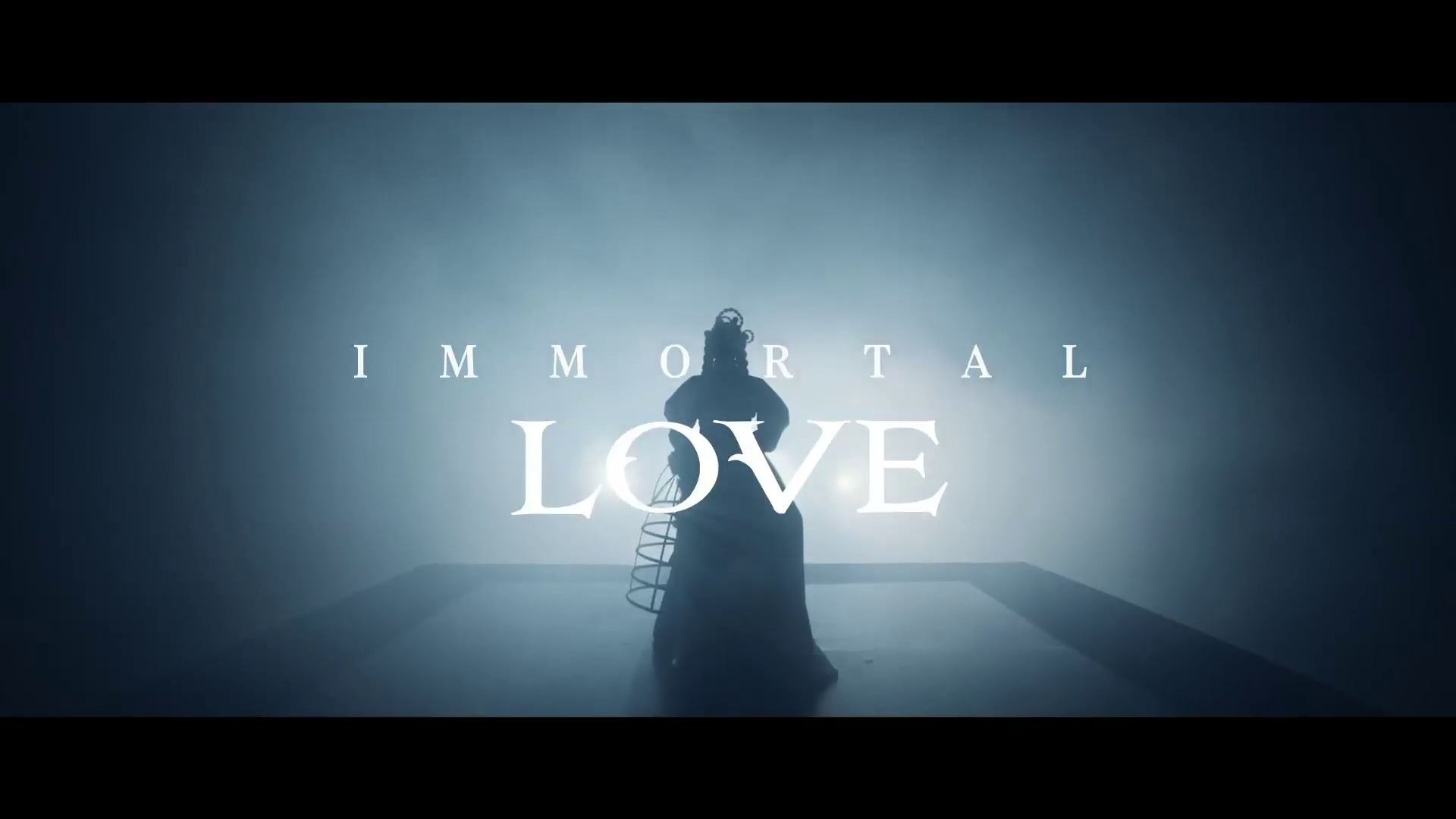 Sylwetka postaci w teatralnej pozie, otoczona metalową konstrukcją, na tle mglistej przestrzeni z napisem 'IMMORTAL LOVE'.