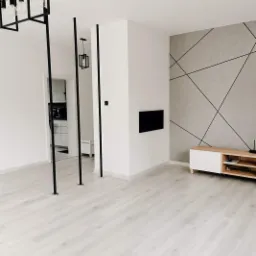 Jasne panele podłogowe w minimalistycznym salonie z szarą ścianą zdobioną geometrycznym wzorem z czarnych linii, czarne pionowe elementy dekoracyjne i nowoczesna lampa wisząca.