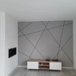 Szara ściana z dekoracyjnymi czarnymi liniami, biała szafka RTV z drewnianym blatem i czarny wkład kominkowy w białej ścianie.