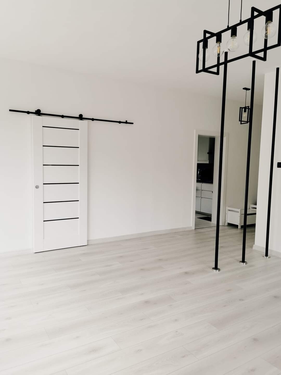Nowoczesne wnętrze z przesuwnymi białymi drzwiami z czarnymi akcentami, drewnianą podłogą, minimalistycznym oświetleniem i otwartym przejściem do innego pomieszczenia.