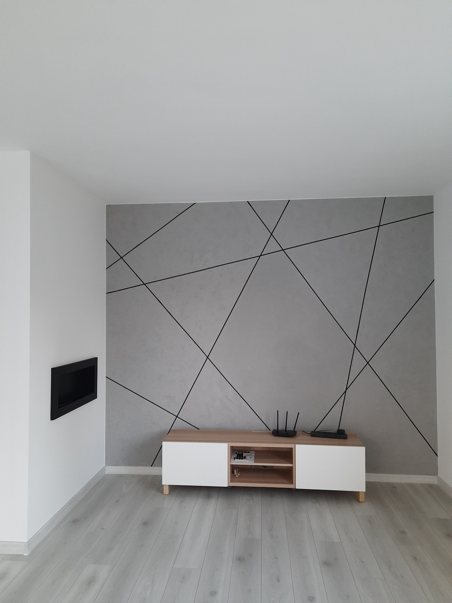 Szara ściana z dekoracyjnymi czarnymi liniami, biała szafka RTV z drewnianym blatem i czarny wkład kominkowy w białej ścianie.