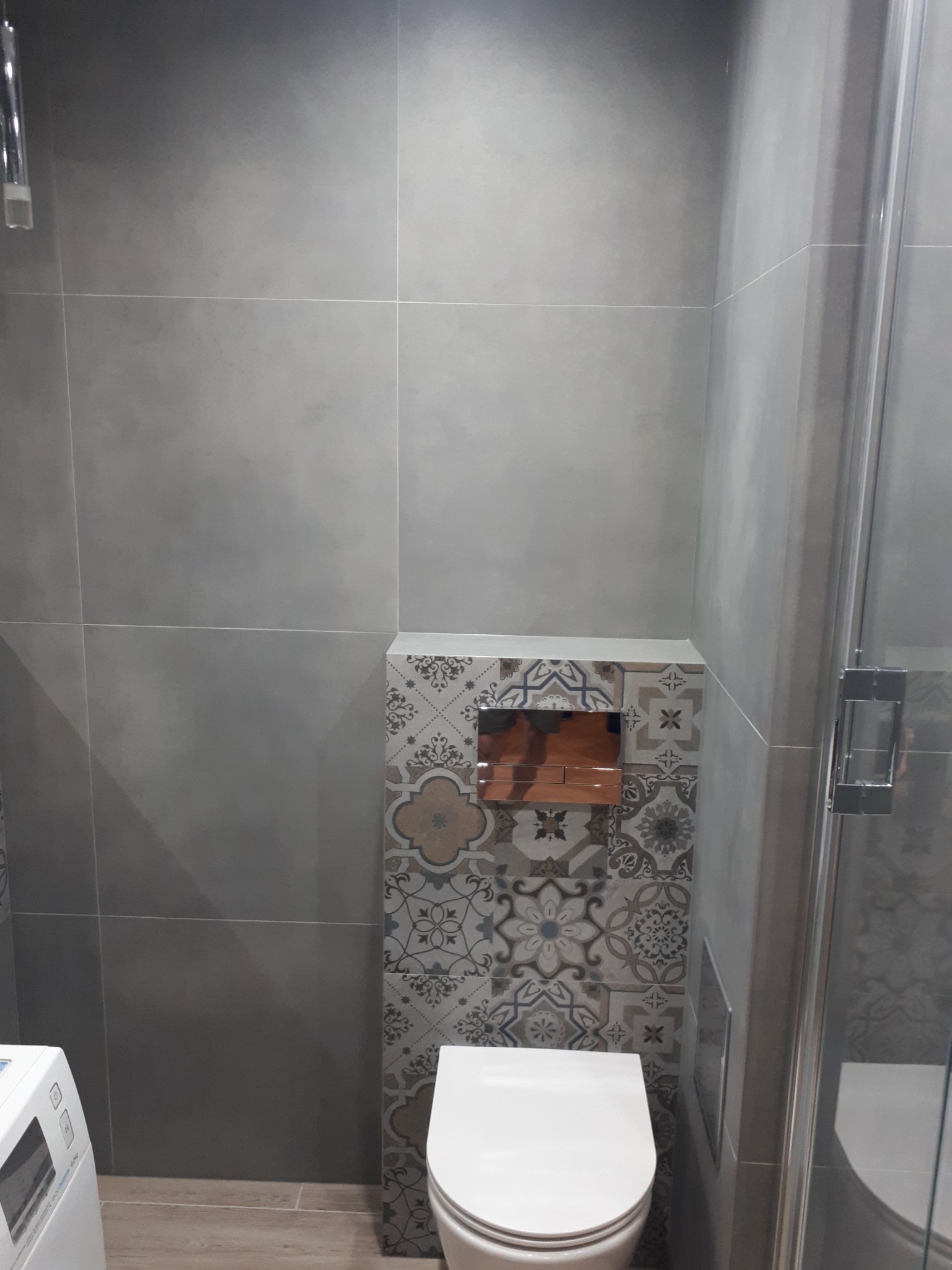 Szara łazienka z płytkami imitującymi beton na ścianach, toaleta podwieszana z mozaiką wzorzystą i kabina prysznicowa ze srebrnymi profilami.