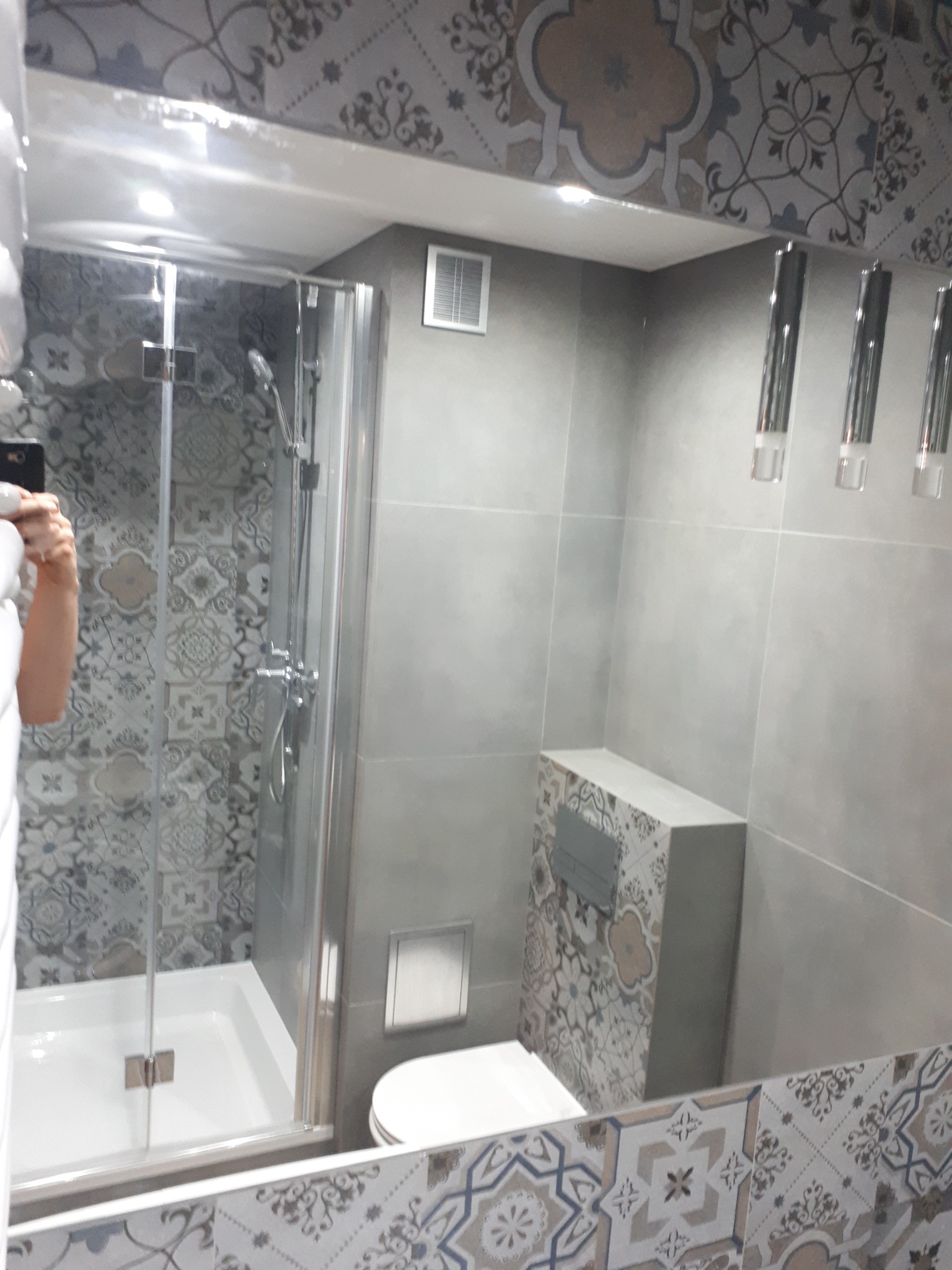 Wykończona łazienka z kabiną prysznicową, toaletą w zabudowie i wzorzystymi płytkami ceramicznymi w odcieniach szarości i beżu, widoczna w lustrze z ozdobną ramą.