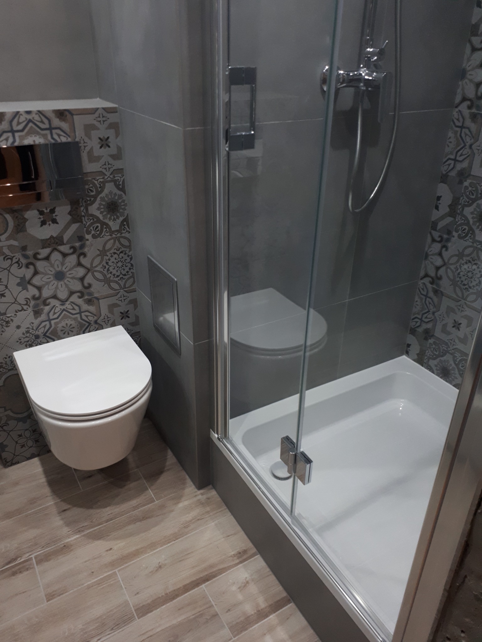 Nowoczesna łazienka z kabiną prysznicową, wiszącą toaletą i dekoracyjnymi płytkami na ścianie, widoczne drewnopodobne płytki podłogowe.