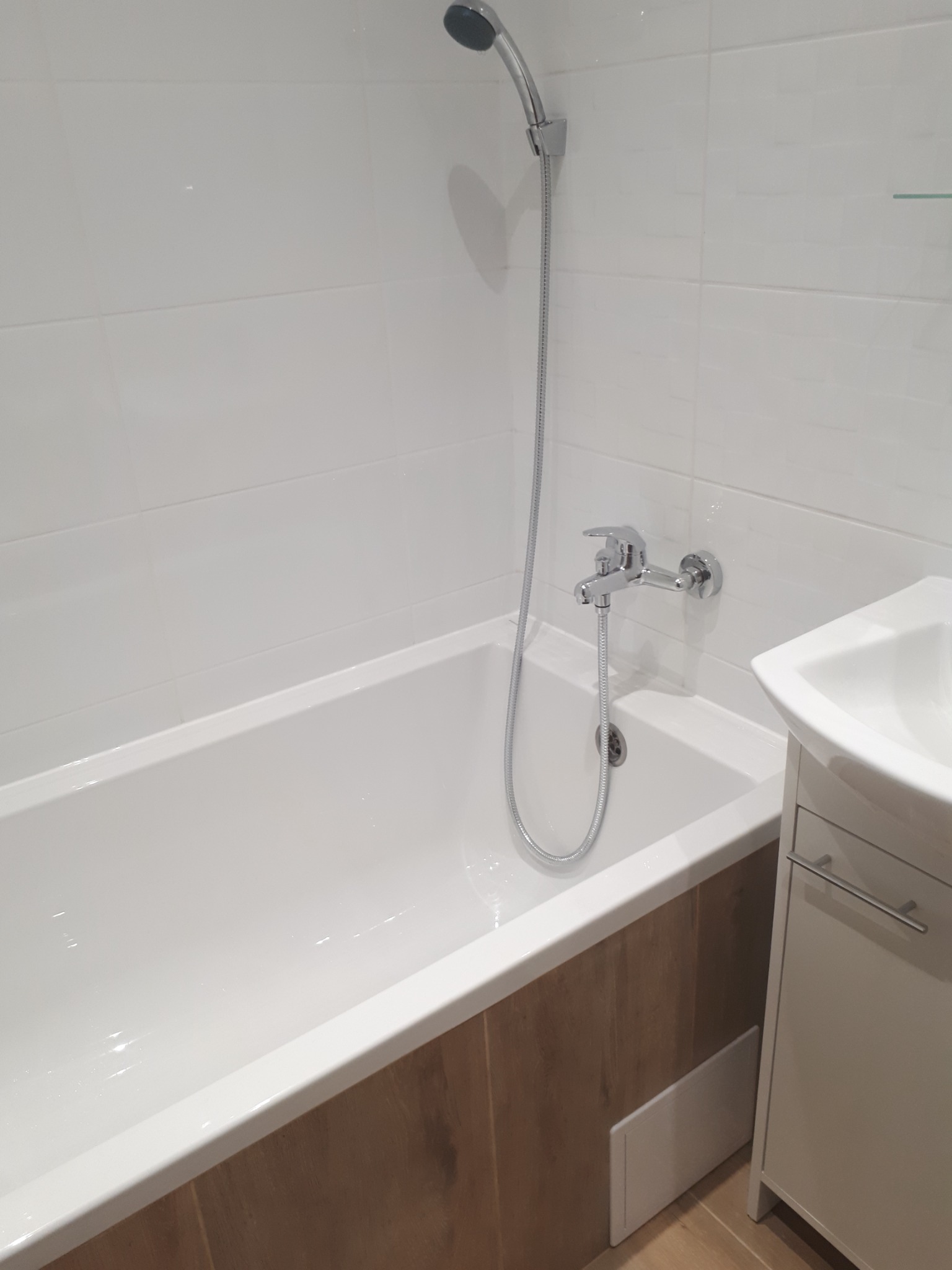 Biała wanna z chromowaną baterią i słuchawką prysznicową na tle białych płytek, z drewnopodobną okładziną i białą szafką umywalkową z boku.