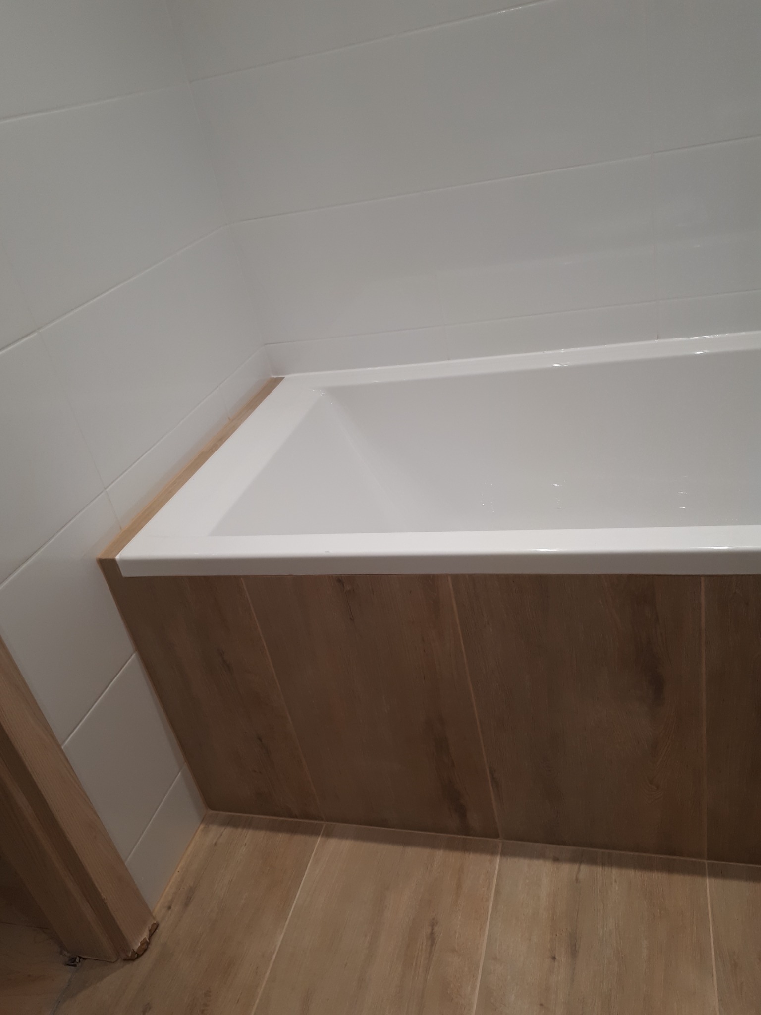 Biała wanna obudowana panelami imitującymi drewno, białe płytki na ścianach i podłoga z płytek drewnopodobnych, widoczny fragment ościeżnicy drzwi.