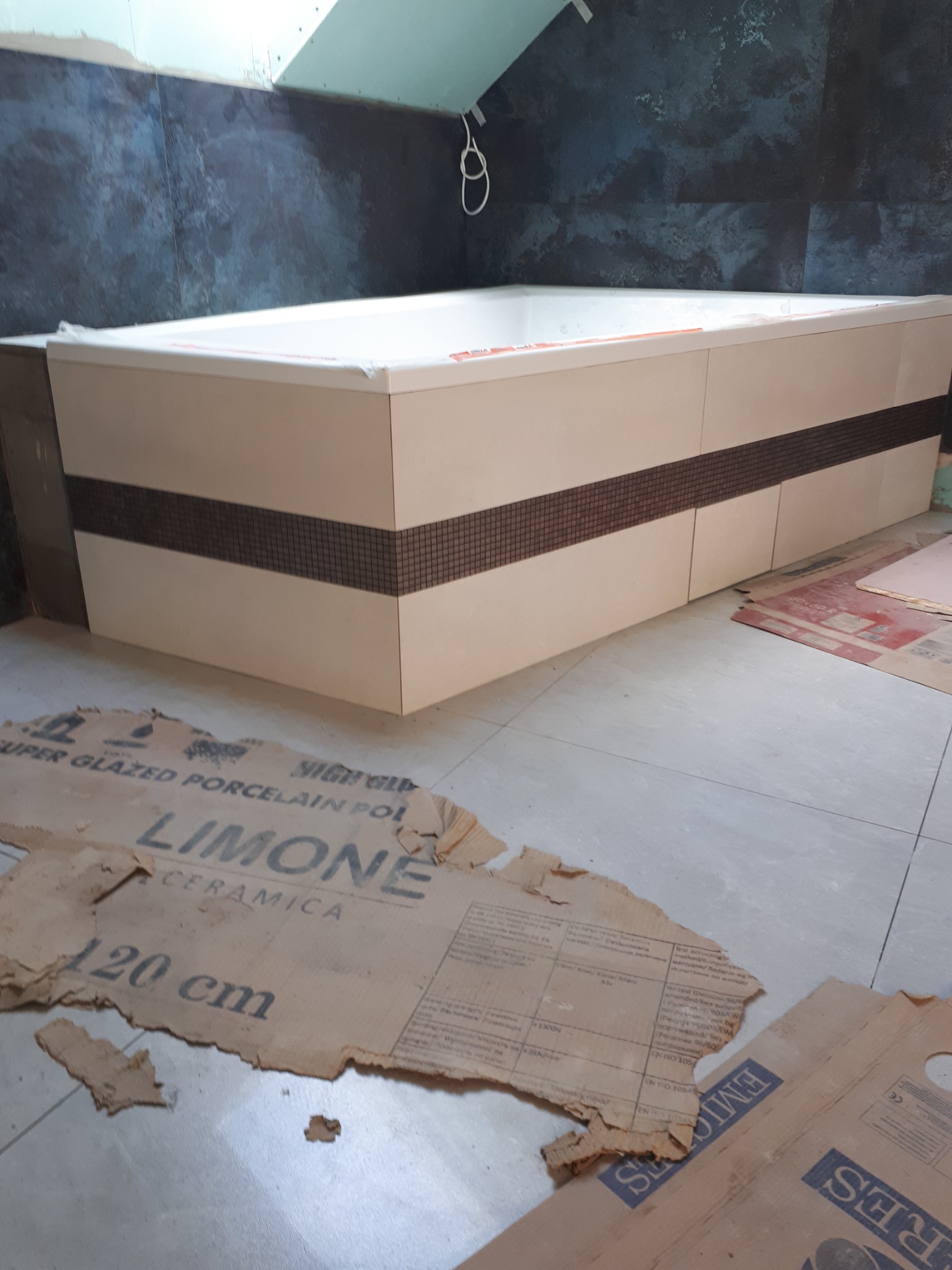 Wanna obudowana płytami z dekoracyjnym pasem mozaiki, widok na fragment łazienki w trakcie prac wykończeniowych, na podłodze kartony zabezpieczające.