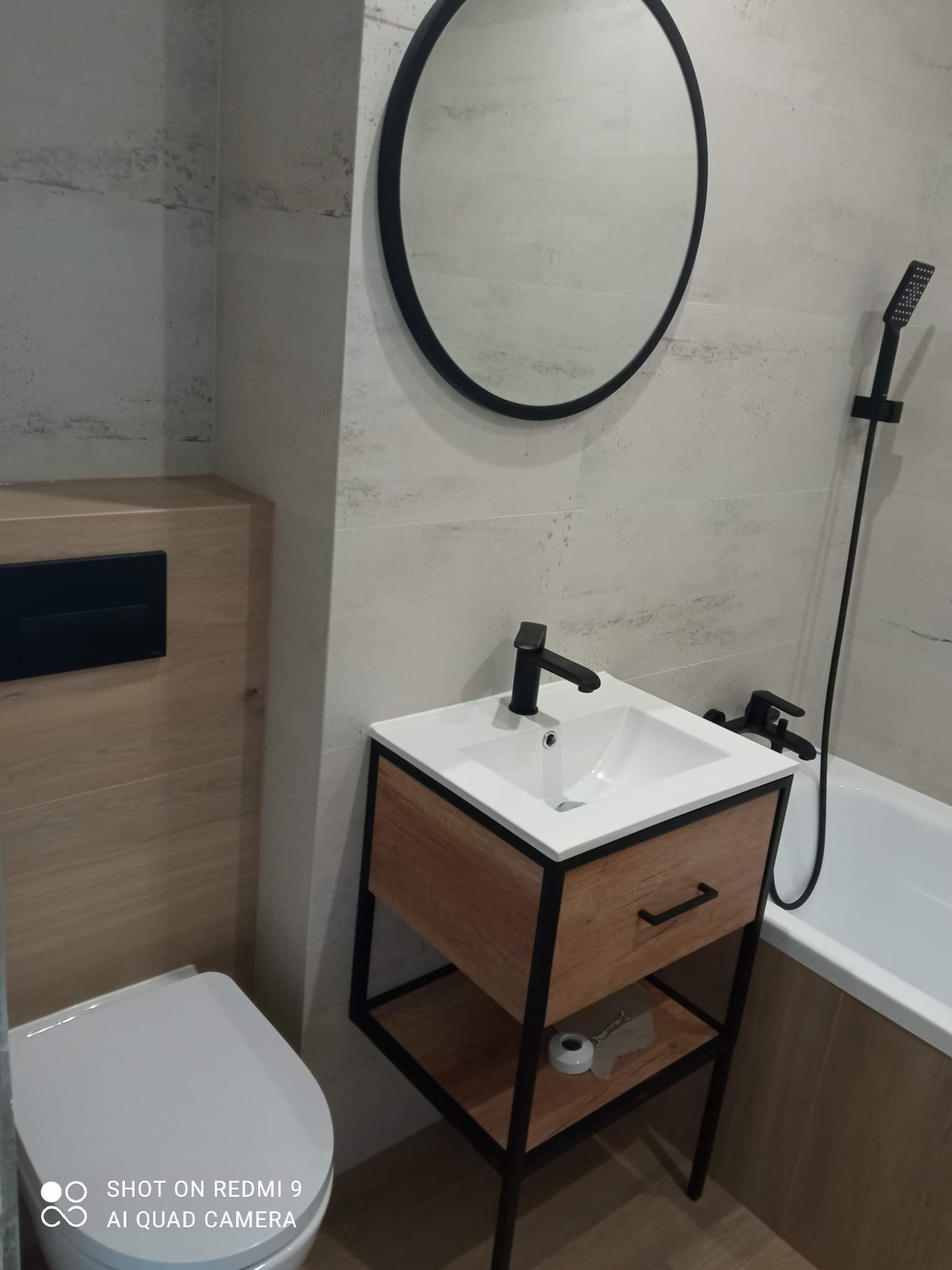 Nowoczesna łazienka z umywalką na drewnianym stelażu z czarnymi elementami, okrągłe lustro w czarnej ramie, biała toaleta i wanna z czarną armaturą.