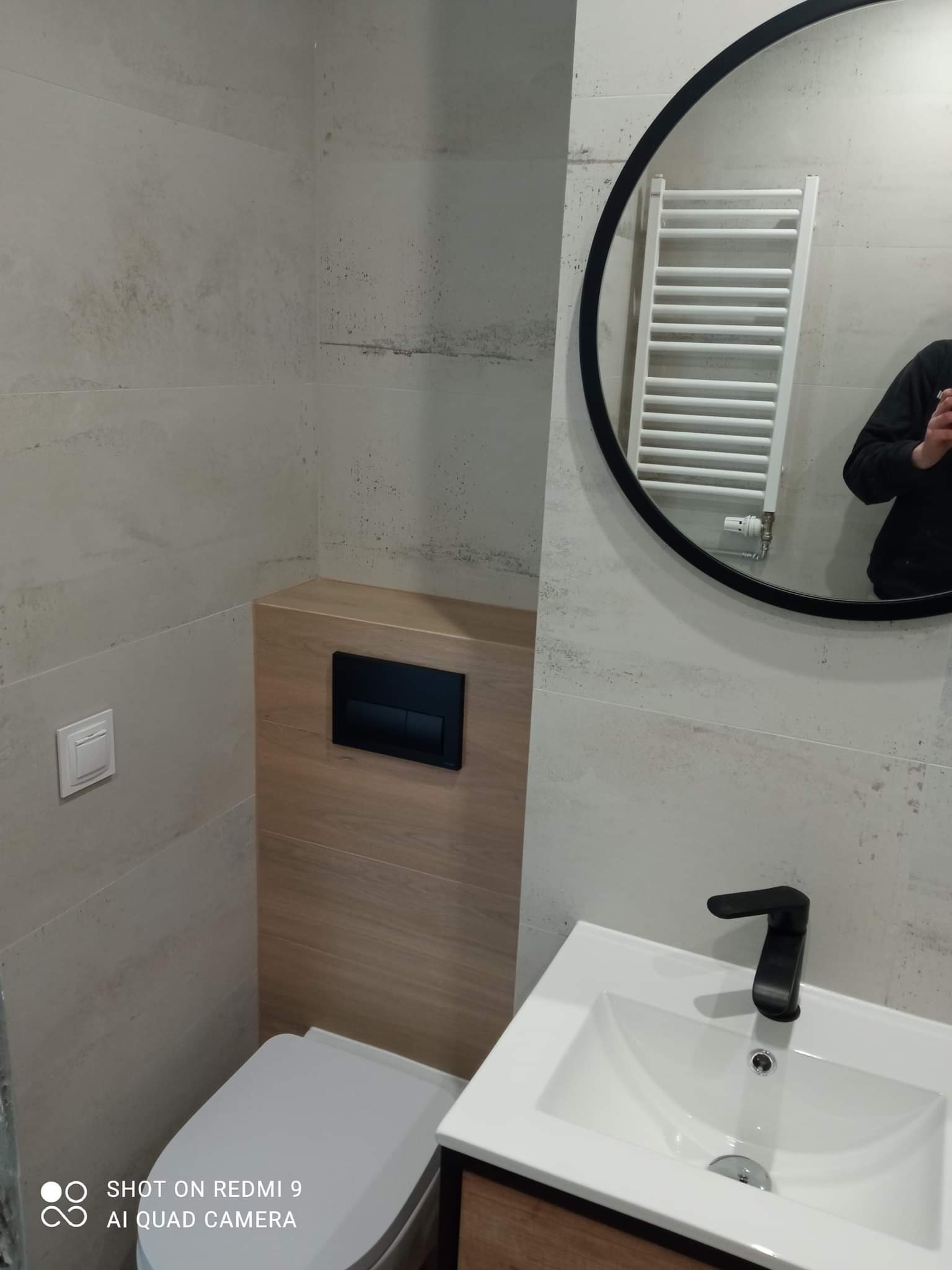 Nowoczesna łazienka z szarymi płytkami imitującymi kamień, podwieszana toaleta z drewnianą obudową, okrągłe lustro z czarną ramą odbijające biały grzejnik drabinkowy i biała umywalka z czarną baterią.