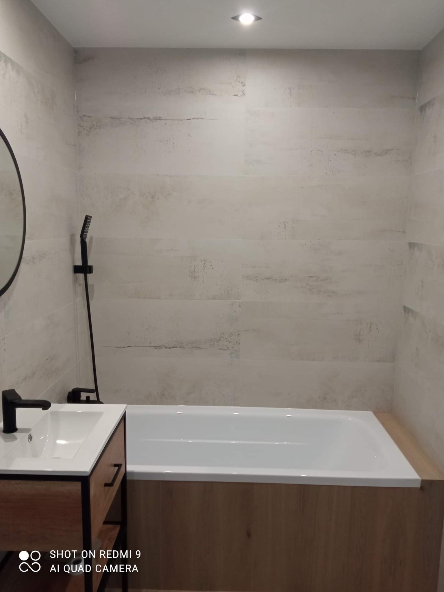 Nowoczesna łazienka z wanną obudowaną drewnopodobną płytą, umywalką z szafką w stylu loft i czarną armaturą na tle płytek imitujących beton.