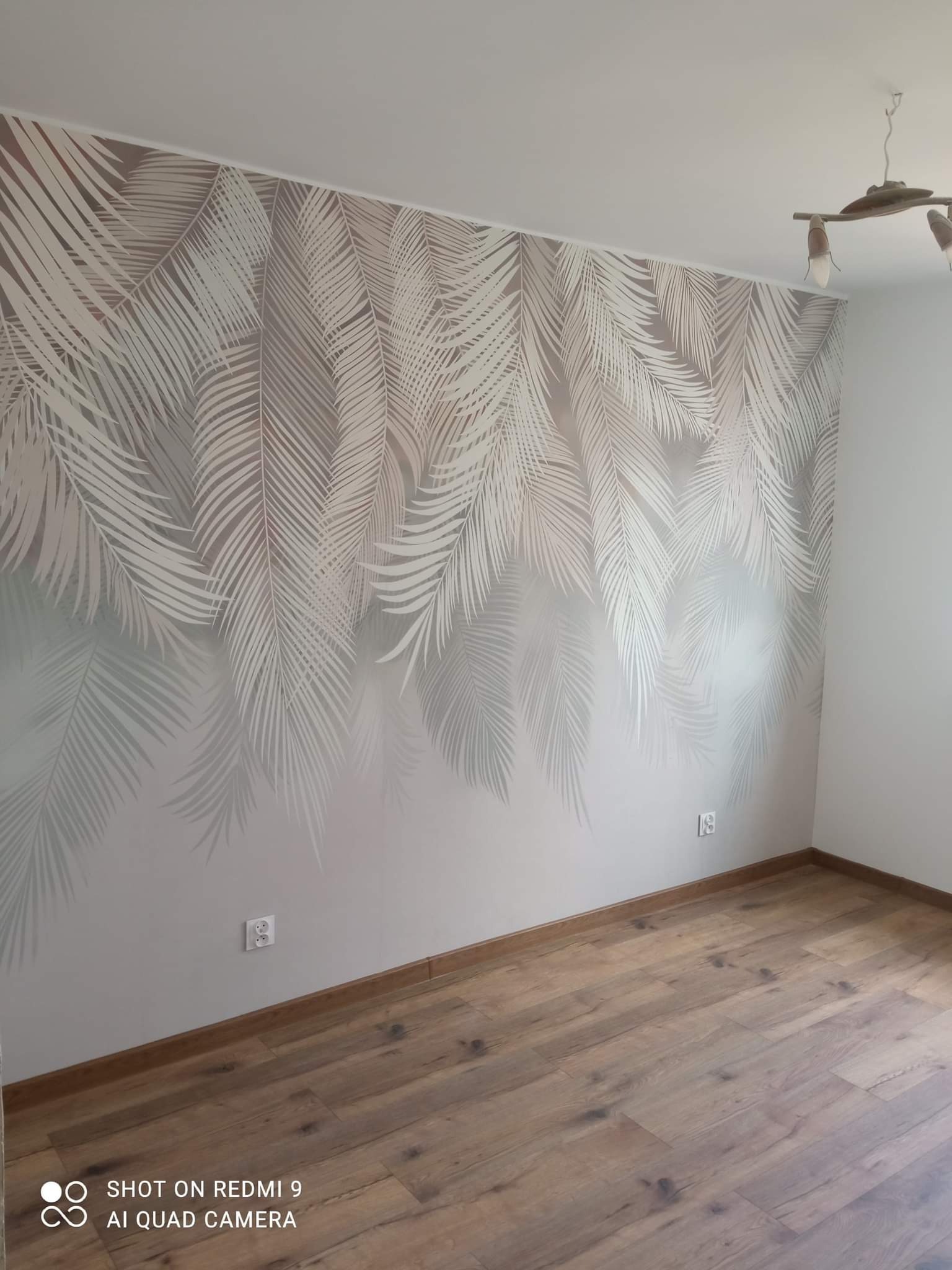 Pokój w Grudziądzu po malowaniu, ściana z motywem liści palmowych w odcieniach szarości, drewniana podłoga, widoczne gniazdka elektryczne i nowoczesna lampa sufitowa.