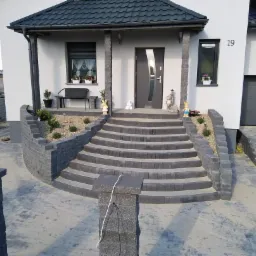 Nowoczesny dom z wejściem wyłożonym szarą kostką brukową, schody z kostki prowadzą do drzwi wejściowych, ogródek z niskimi roślinami, numer domu 19 nad oknem, widoczna boczna ściana domu z bramą...