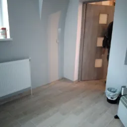 Jasne płytki podłogowe imitujące drewno ułożone w nowym pomieszczeniu z łukowym sufitem, widoczny biały grzejnik, drzwi z szybkami i fragment osoby w tle.