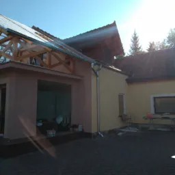 Dom jednorodzinny w trakcie prac elewacyjnych i dekarskich, widoczne rusztowania, częściowo otynkowane ściany, nowe rynny i drewniana konstrukcja dachu, narzędzia na zewnątrz.