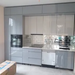 Jasnoszara, nowoczesna zabudowa kuchenna z piekarnikiem, płytą indukcyjną i zmywarką, uzupełniona o lustrzane płytki nad blatem, z kartonem na podłodze.