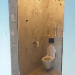 Wnętrze wykończonej łazienki z podwieszaną toaletą, marmurowymi płytkami, ozdobnymi świecznikami i kryształowym żyrandolem, widok zza surowej ościeżnicy.