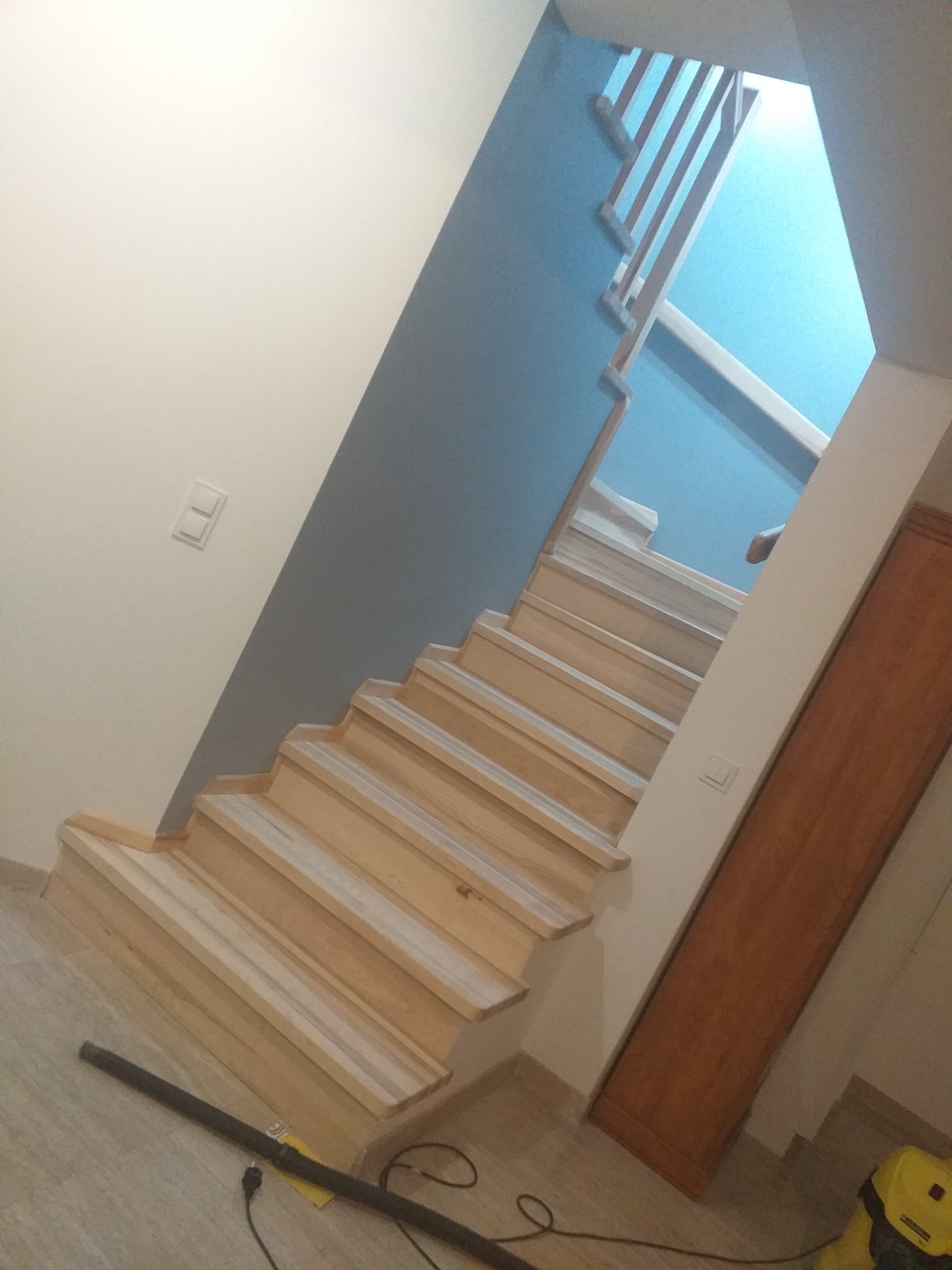 Jasne drewniane schody z balustradą na niebieskiej ścianie, widoczne w trakcie montażu, z rurą od odkurzacza na podłodze.