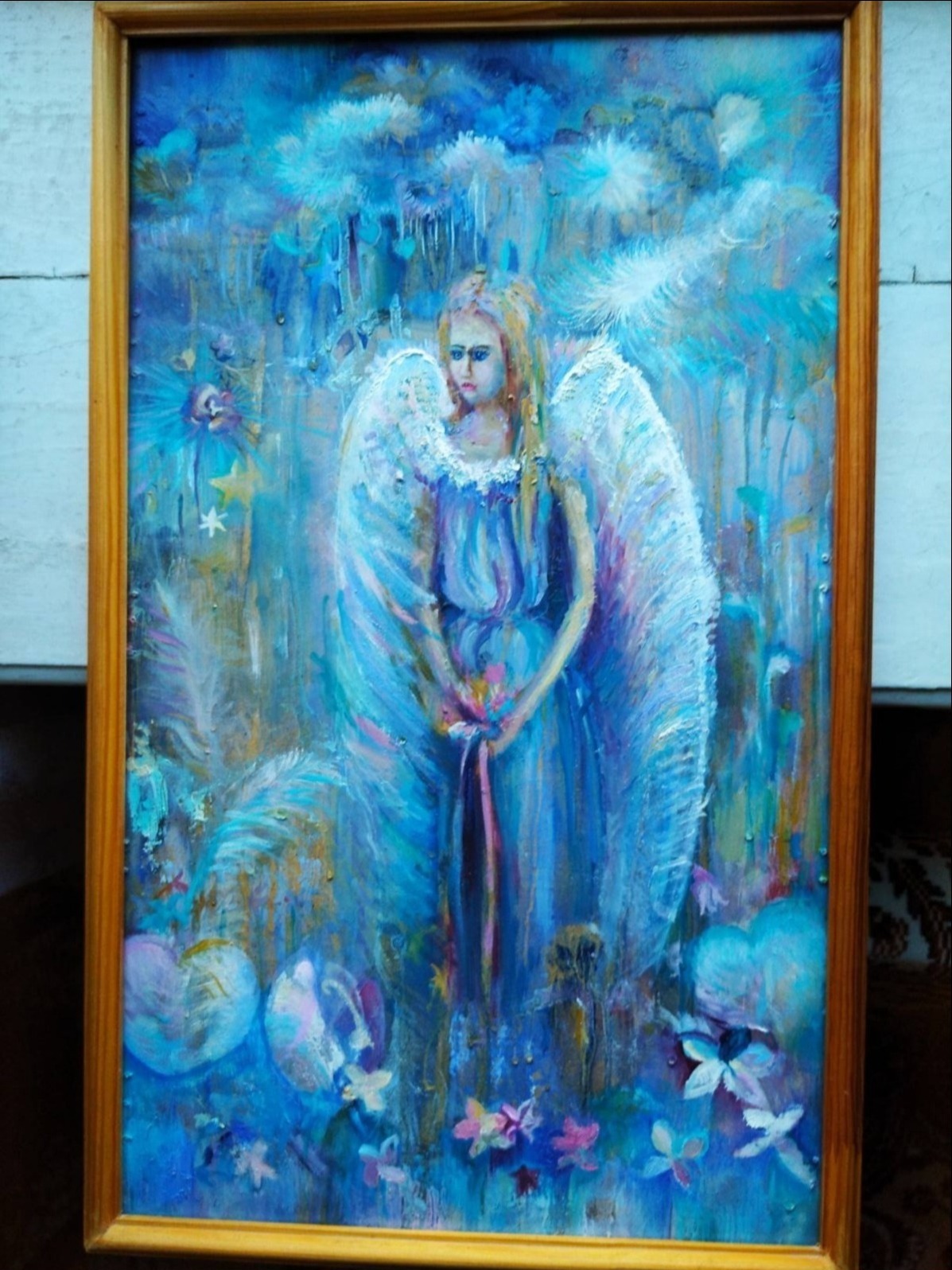 Obraz olejny w drewnianej ramie przedstawiający anioła w błękitnej sukni z białymi skrzydłami na tle abstrakcyjnego, błękitnego krajobrazu z kwiatami.