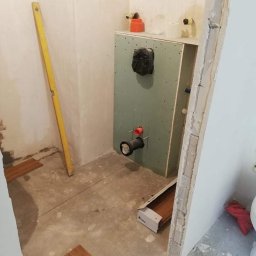 SzWykończenia - Przygotowanie do montażu podwieszanego WC: stelaż obudowany płytą gipsowo-kartonową, widoczne rury instalacyjne, poziomica oparta o ścianę, fragment podłogi z rozłożonymi panelami imitującymi drewno.