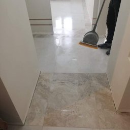 SzWykończenia - Układanie jasnych, marmurkowych płytek podłogowych w korytarzu, widoczny pracownik sprzątający szczotką z szufelką, przejście do kolejnego pomieszczenia z białymi ścianami i jasną podłogą.