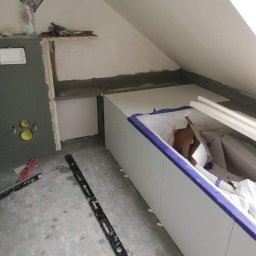 SzWykończenia - Poddasze w trakcie wykańczania: zabudowa z płyt gipsowo-kartonowych wzdłuż ściany, widoczne profile, poziomica na podłodze, przygotowane miejsce na umywalkę z instalacją hydrauliczną.