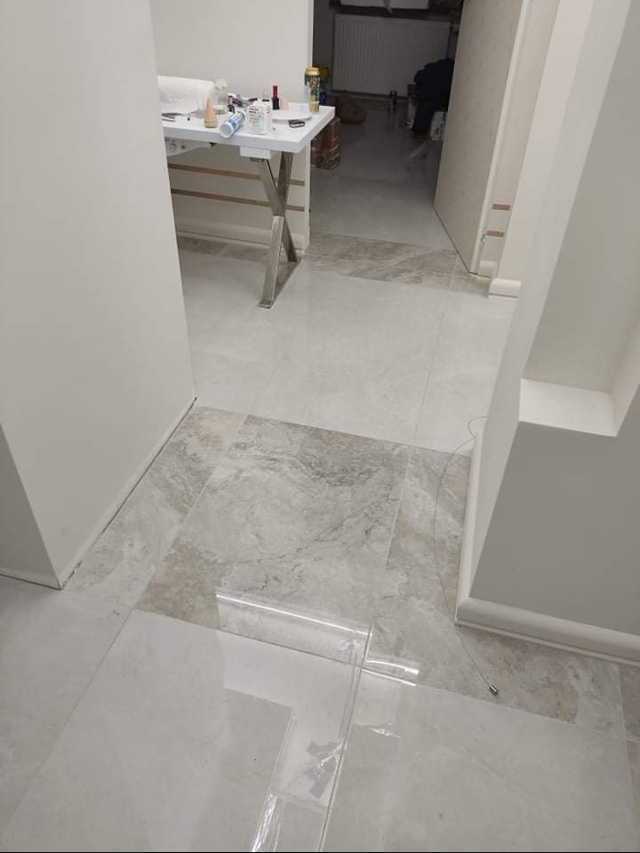 Ułożone szare płytki podłogowe w korytarzu, fragment zabezpieczony folią ochronną, w tle widoczny stół z narzędziami i materiałami budowlanymi.