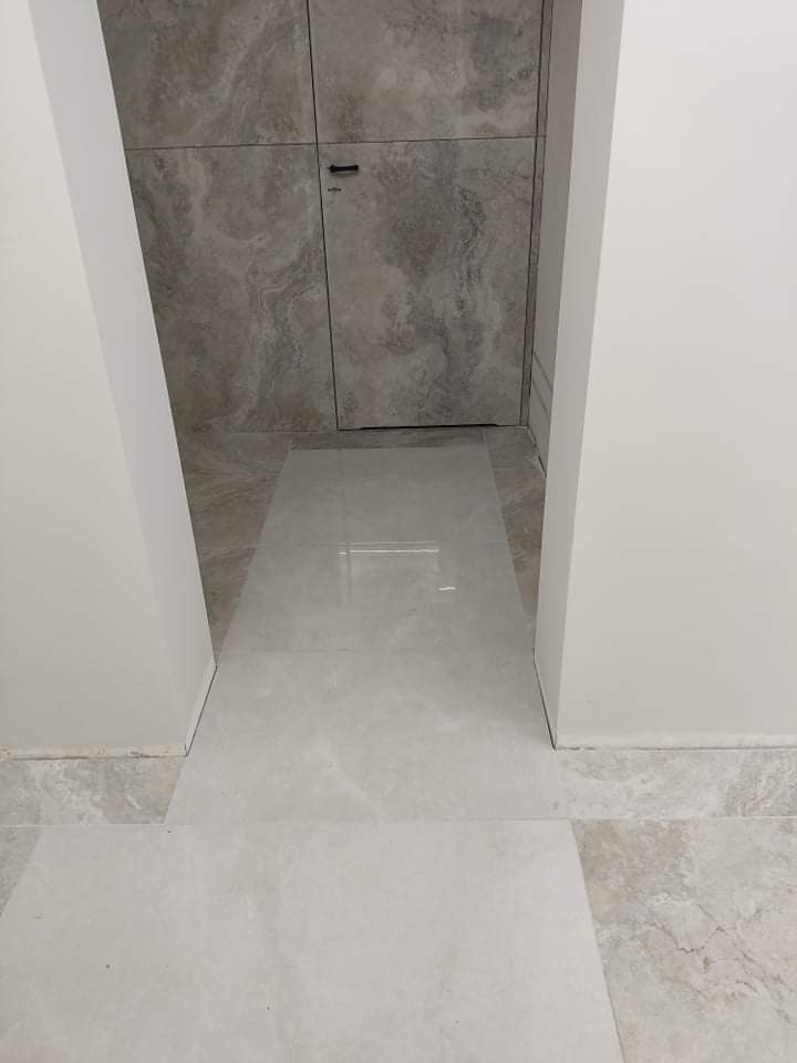 Wykończona podłoga z jasnych, marmurkowych płytek, prowadząca do ściany z ukrytymi drzwiami wykończonymi szarymi płytkami imitującymi kamień, z widoczną małą klamką.