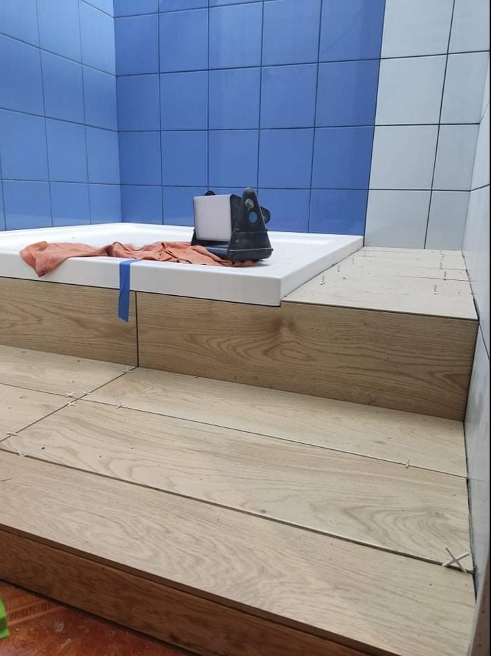 Montaż płytek imitujących drewno na podwyższeniu w łazience, z użyciem krzyżyków dystansowych i narzędzia do cięcia płytek na wannie.