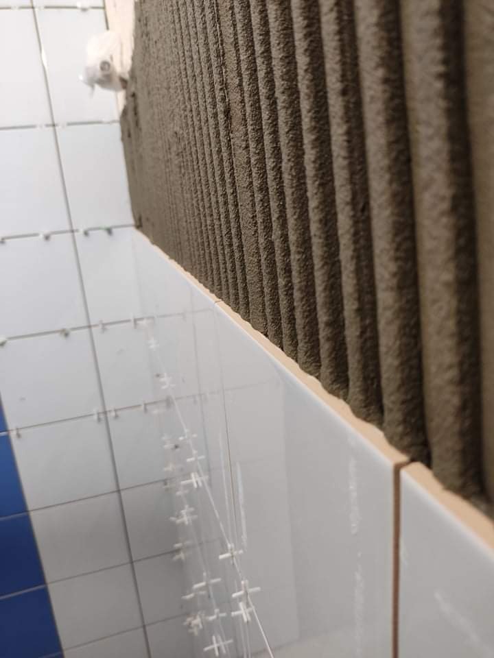 Świeżo nałożona warstwa kleju z wyraźnymi rowkami na ścianie, obok ułożone jasne płytki z krzyżykami dystansowymi, widok z bliska.