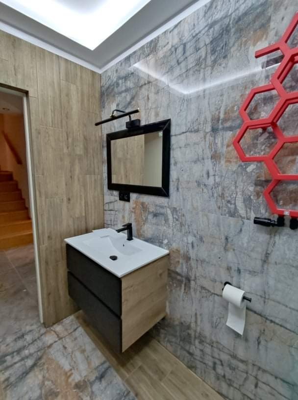Nowoczesna łazienka z umywalką nablatową na szafce w kolorze drewna i grafitu, lustro z oświetleniem LED, ściana z płytek imitujących kamień z podświetleniem i czerwonym grzejnikiem dekoracyjnym...