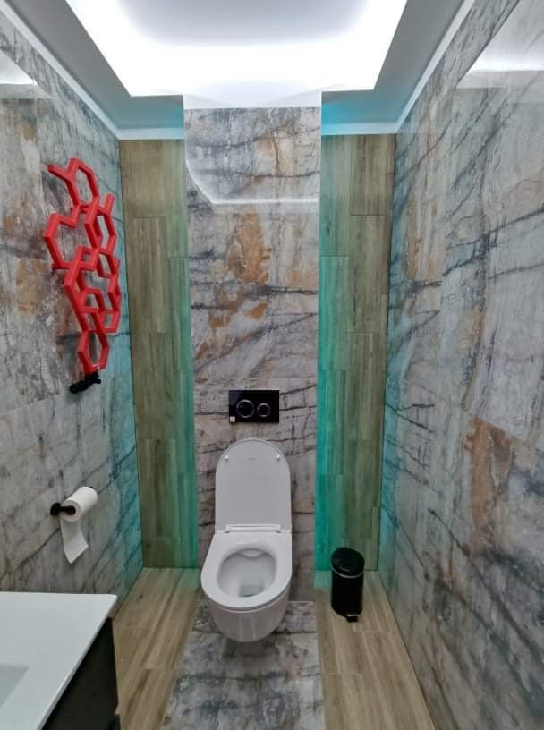 Wykończona łazienka z marmurowymi płytkami, podświetlanym sufitem, toaletą z czarnym panelem sterowania i dekoracyjnym czerwonym elementem na ścianie.