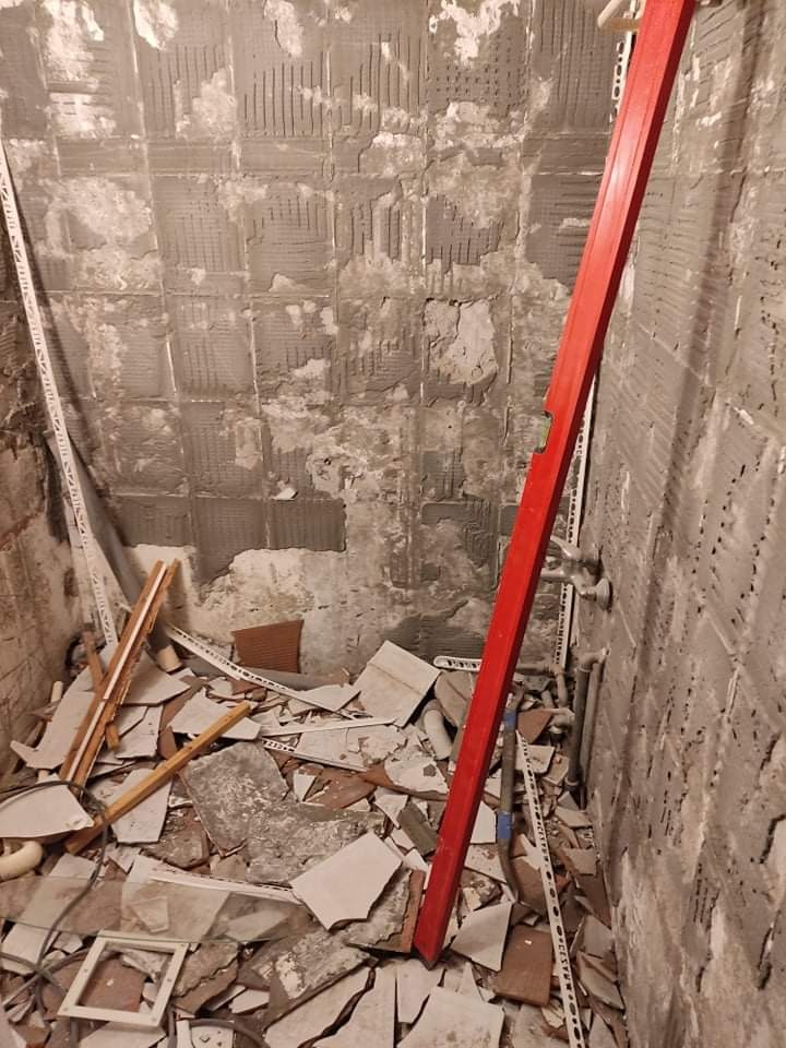Demontaż starej glazury w łazience, widoczne resztki kleju na ścianach, gruz z potłuczonych płytek na podłodze i czerwona poziomica oparta o ścianę.