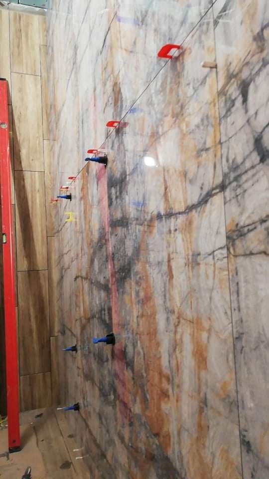 Ściana w trakcie układania płytek imitujących marmur, z widocznymi klipsami i klinami poziomującymi w kolorach czerwonym, niebieskim i żółtym, oraz pionową poziomicą przy sąsiedniej ścianie...