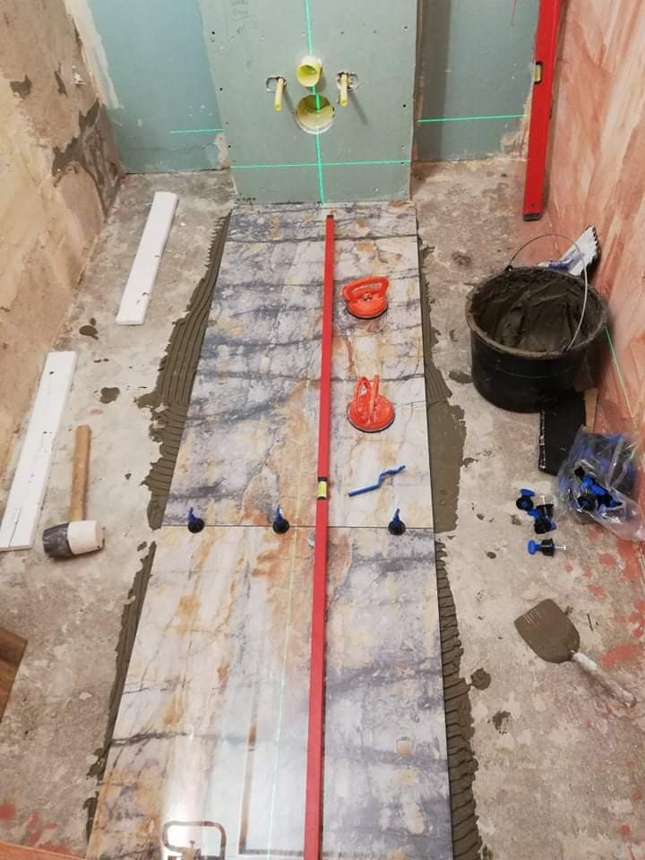 Układanie płytek podłogowych w łazience, widoczny laser krzyżowy, przyssawki do płytek, poziomica i wiadro z zaprawą.