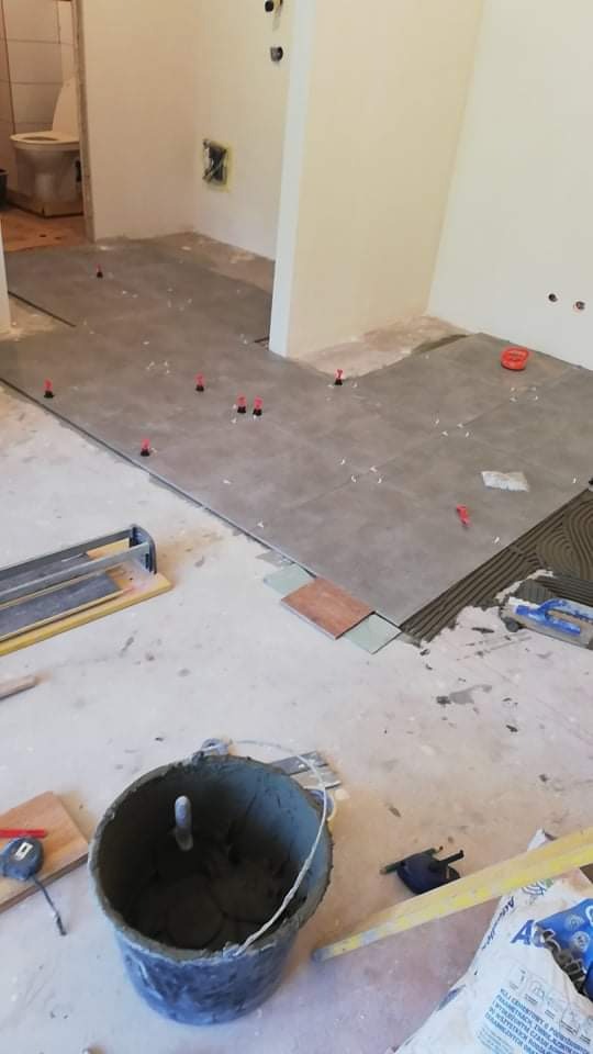 Układanie szarych płytek podłogowych w łazience z widocznymi krzyżykami dystansowymi, narzędziami glazurniczymi i wiaderkiem z zaprawą.