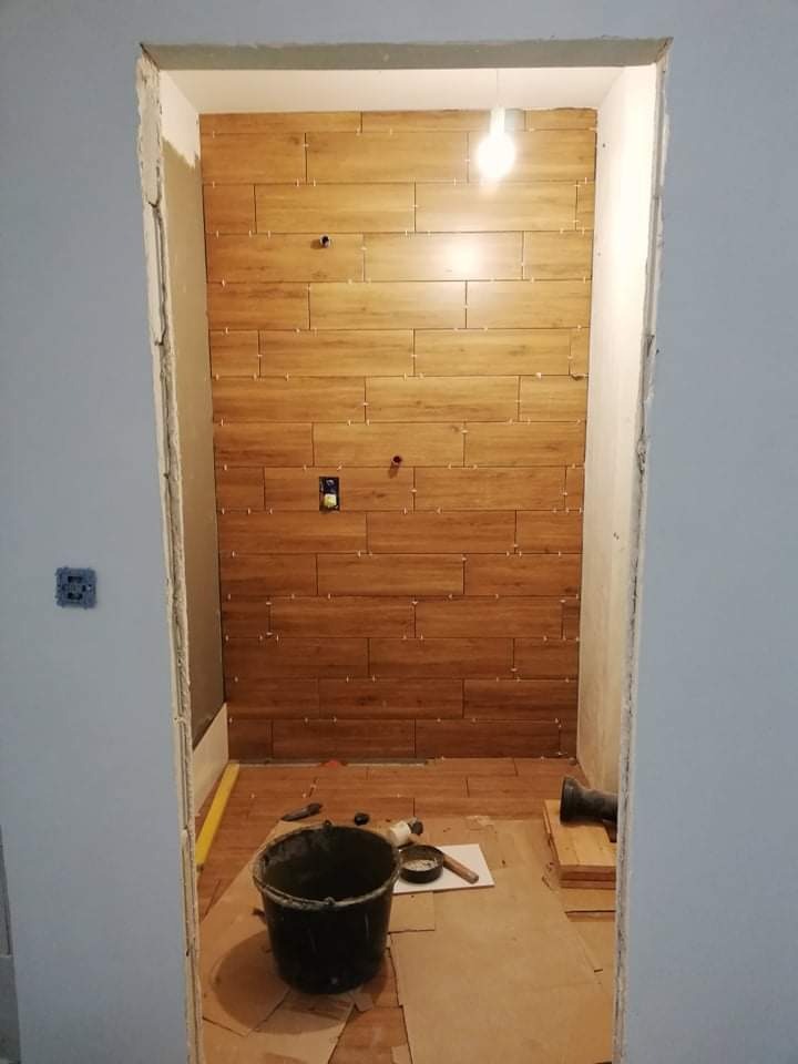 Ściana łazienki w trakcie układania płytek imitujących drewno, widoczne krzyżyki dystansowe, puszka elektryczna i przygotowane narzędzia pracy na podłodze zabezpieczonej kartonem.