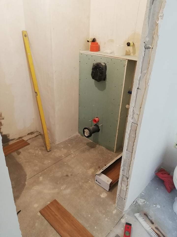 Przygotowanie do montażu podwieszanego WC: stelaż obudowany płytą gipsowo-kartonową, widoczne rury instalacyjne, poziomica oparta o ścianę, fragment podłogi z rozłożonymi panelami imitującymi drewno.